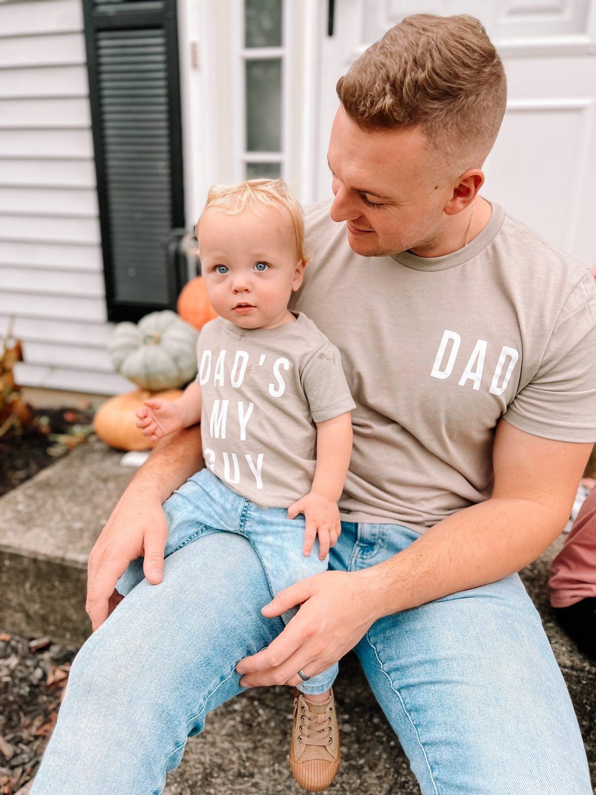 A dad and son wearing a set of brown “Dad’s my guy” t-shirts