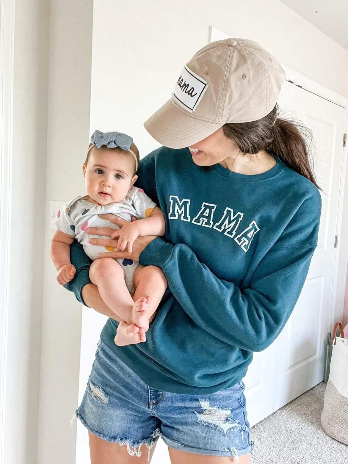 A mom holding her baby and wearing the Varsity Mama Teal Pullover