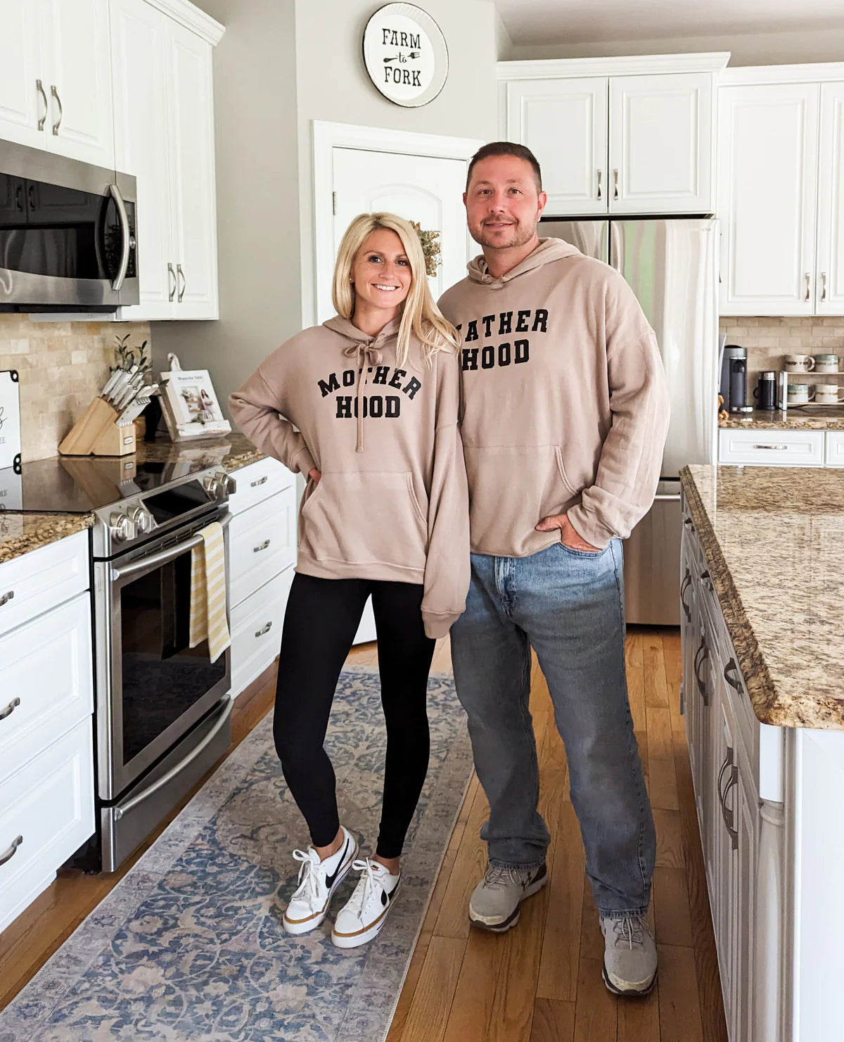 A mom and dad wearing a set of brown hoodies that say “motherhood” and “fatherhood”